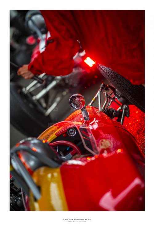 Voiture de course rouge en pré-grille du grand prix historique de Pau, un assistant protège le pilote de la pluie